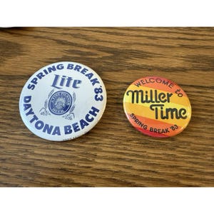 May include: Two circular pinback buttons. The larger white button features blue text reading SPRING BREAK '83 DAYTONA BEACH and a Lite beer logo. The smaller button has horizontal orange and yellow stripes with black text reading WELCOME TO MILLER TIME SPRING BREAK '83.