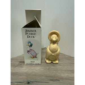 May include: A cream-colored duck-shaped soap figurine standing next to its original white cardboard box. The box features an illustration of Jemima Puddle-Duck, the text "Jemima Puddle-Duck SOAP", and the weight "net wt 105 g 3.7 oz".