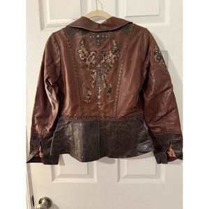 May include: A brown leather jacket featuring a dark brown peplum hem with a textured finish. The back panel displays intricate decorative stitching, metallic stud accents, and a central patterned inlay design. It has long sleeves with cuffs.
