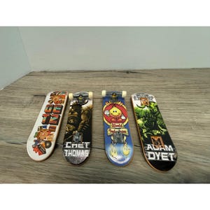 May include: Four finger skateboards with distinct graphic designs. The first features colorful text and illustrations, the second shows a dark creature with the name Chet Thomas, the third displays a yellow flame character, and the fourth depicts a green warrior with the name Adam Dyet.