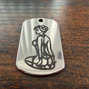 May include: A silver-toned metal dog tag pendant with a rounded rectangular shape and a hole at the top. The surface features a black laser-engraved line art illustration of a person with an updo hairstyle, shown from the side while seated.