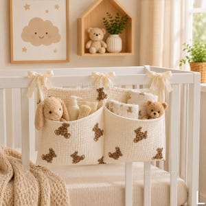 Könnte beinhalten: Ein cremefarbener Stoff-Organizer für das Kinderbett mit zwei tiefen Taschen, verziert mit einem braunen Teddybärenmuster und befestigt an einem weißen Gitterbett mit drei cremefarbenen Schleifen. Die Taschen enthalten einen Plüschhasen und einen Teddy.