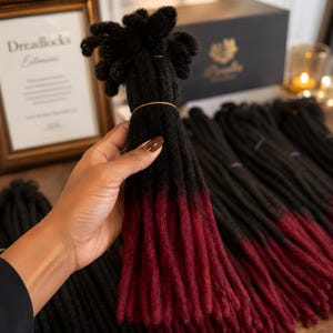 Puede incluir: Un manojo de extensiones de rastas sintéticas con una base negra que se transforma en un color rojo burdeos intenso en las puntas. Los mechones individuales tienen un aspecto texturizado y mate, sujetos con una fina banda de color dorado.