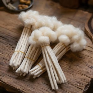 Peut inclure: Trois paquets de laine cardée blanc cassé, chacun présentant une touffe de fibres duveteuses non filées au sommet et plusieurs longs brins torsadés maintenus ensemble par un élastique fin de couleur claire près du centre de la longueur.