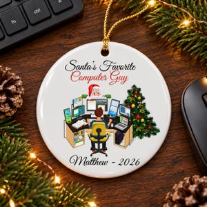 May include: A round white ceramic ornament featuring a graphic of a person sitting at a desk with multiple computer monitors, a Christmas tree, and Santa Claus peeking over the screens. Text reads Santa's Favorite Computer Guy and Matthew - 2026.