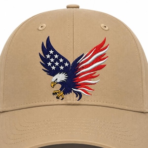 May include: A tan baseball cap featuring an embroidered graphic of an eagle. The eagle's left wing displays a blue field with white stars, while the right wing features red and white stripes. The eagle has a white head, yellow beak, and yellow talons.