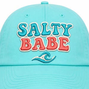 Può includere: Un cappellino da baseball azzurro con il testo ricamato SALTY BABE in uno stile a strati con lettere blu e rosse. Sotto il testo si trova un grafico ricamato di un'onda oceanica stilizzata in filo blu e bianco sul pannello frontale.