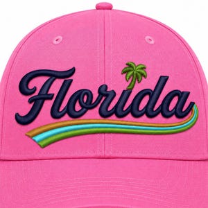 Op de afbeelding: Een felroze honkbalpet met het woord Florida geborduurd in donkerblauw cursief schrift. Boven de tekst staat een klein groen palmboomicoon en onder de tekst bevinden zich drie gebogen lijnen in de kleuren bruin, lichtblauw en limoengroen.