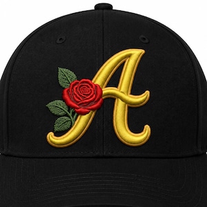 May include: A black baseball cap featuring an embroidered design on the front. The design consists of a large, golden yellow letter A in a cursive font, accented by a detailed red rose with green leaves positioned to the left of the letter.