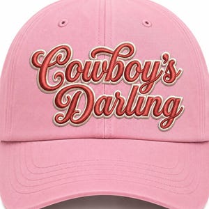 May include: A light pink baseball cap featuring the text Cowboy's Darling embroidered in a bold, red script font with a white outline. The cap has a structured crown, a curved brim, and matching pink stitching details across the front panels.