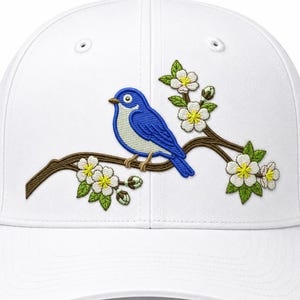 Puede incluir: Una gorra de béisbol blanca con un diseño bordado de un pájaro azul posado en una rama de árbol marrón. La rama está decorada con flores blancas con centros amarillos y hojas verdes, creando un gráfico detallado inspirado en la naturaleza.