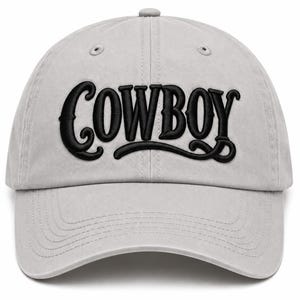 May include: A light gray baseball cap featuring the word COWBOY embroidered in bold black lettering across the front panels. The cap has a structured crown, a curved brim with stitched detailing, and eyelets for ventilation on the top panels.