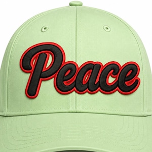 May include: A light green baseball cap featuring the word Peace embroidered in black with a bold red outline across the front panels. The cap has a structured crown, a curved brim, and stitched eyelets for ventilation on the top panels.