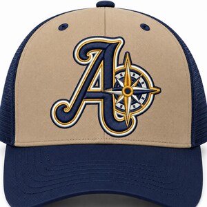 May include: A tan and navy blue baseball cap featuring an embroidered navy blue letter A with a gold and white compass rose design integrated into the right side of the letter. The cap has a solid tan front panel and navy blue mesh back panels.