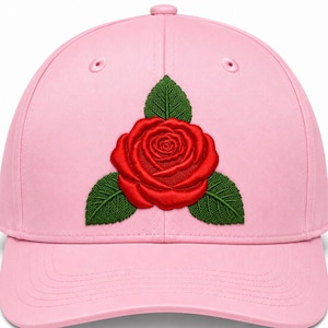 Puede incluir: Una gorra de béisbol de color rosa claro con un bordado detallado y centrado de una vibrante flor de rosa roja rodeada por tres hojas de color verde oscuro. La gorra presenta una corona estructurada con ojales rosas a juego.