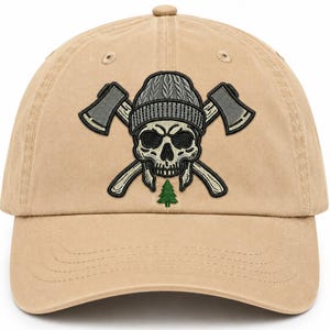May include: A tan baseball cap featuring an embroidered graphic of a skull wearing a grey knit beanie, positioned in front of two crossed axes. Below the skull is a small, green embroidered pine tree, centred on the front panel of the cap.