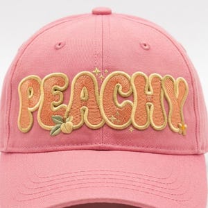 May include: A pink baseball cap featuring the word PEACHY embroidered in orange and gold thread across the front. The design includes small embroidered peach fruit, green leaves, and tiny star accents integrated into the lettering.