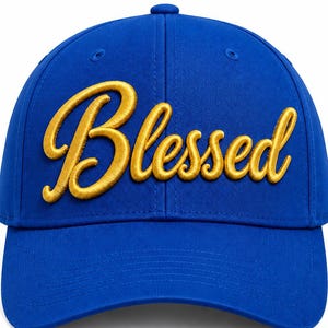 May include: A vibrant royal blue baseball cap featuring the word Blessed embroidered in a raised, metallic gold script font across the front panels. The cap has a structured crown, a curved brim, and matching blue stitching throughout the design.