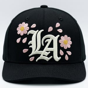 May include: A black baseball cap featuring the letters LA embroidered in a white gothic font. The design is accented with embroidered pink flowers with yellow centers and scattered pink petals surrounding the central lettering on the front panel.