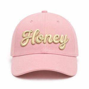 May include: A light pink baseball cap featuring the word Honey embroidered in a raised, metallic gold script font across the front panels. The cap has a structured crown, a curved brim, and matching pink stitching details throughout the design.