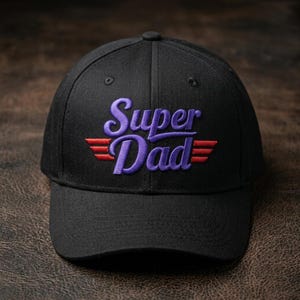 May include: A black baseball cap featuring embroidered text that reads Super Dad in a purple script font. The design includes two sets of three horizontal red lines extending from the sides of the word Dad, creating a wing-like graphic element.