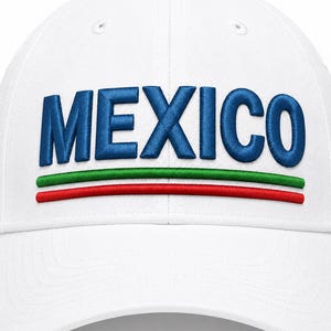 Puede incluir: Una gorra de béisbol blanca con la palabra MEXICO bordada en letras mayúsculas de color azul intenso en la parte frontal. Debajo del texto hay dos líneas bordadas paralelas, una verde y otra roja, que siguen la curva de las letras.