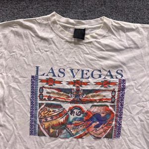 Könnte beinhalten: Ein weißes kurzärmeliges T-Shirt mit dem Schriftzug LAS VEGAS in blauen Buchstaben über einem Grafikdruck. Das Design zeigt einen zentralen Adler, zwei gemusterte Töpfergefäße und geometrische Ränder in Rot-, Blau- und Orangetönen.
