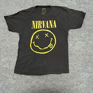 May include: A black short-sleeve t-shirt featuring the yellow Nirvana band logo text and a large yellow smiley face graphic with X-shaped eyes and a tongue sticking out on the front. The inner neck label also displays the Nirvana smiley logo.