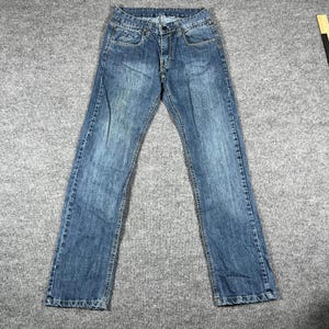 May include: A pair of blue denim jeans featuring a classic five-pocket design, a button and zipper fly closure, belt loops, and a medium wash finish with subtle fading on the thighs and knees, presented with a visible yellow measuring tool.