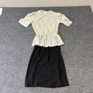 May include: A two-piece outfit consisting of a cream-colored blouse with black polka dots, short puff sleeves, and a peplum waist, paired with a solid black knee-length skirt. The blouse features a round neckline with small black buttons on the shoulder.