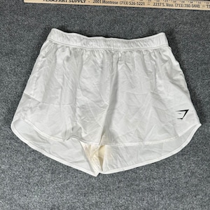 May include: White athletic shorts with an elastic waistband and a black logo on the lower right leg. The shorts are positioned below a ruler that shows measurements in inches, with the visible portion ranging from approximately 15 to 24 inches.