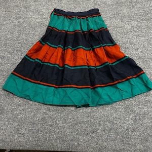 May include: A pleated midi skirt featuring horizontal stripes in dark navy blue, vibrant orange, and bright teal green. The skirt has a fitted waistband and a flared silhouette that extends to a wide hemline with a solid teal green band.