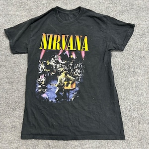 May include: A black short-sleeve t-shirt featuring the word NIRVANA in bold yellow letters at the top. Below the text is a graphic print of a band performing on stage with a musician playing an acoustic guitar in a dimly lit, purple-toned setting.