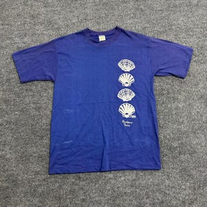 Puede incluir: Una camiseta de manga corta color azul real con una columna vertical de cuatro ilustraciones de conchas marinas blancas en el lado derecho. Debajo de las conchas, el texto Rockport Texas aparece en letra cursiva blanca. Tiene cuello redondo.