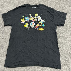 May include: A black short-sleeve t-shirt featuring a colorful graphic print of various cartoon characters floating in space among stars and planets. The Nickelodeon logo is printed in orange at the bottom right of the graphic design.