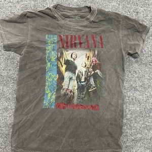 May include: A charcoal gray short-sleeve t-shirt featuring a graphic print of the band Nirvana. The design includes the text NIRVANA in red, a vertical blue textured stripe, and the word NEVERMIND in red at the bottom below the band members.