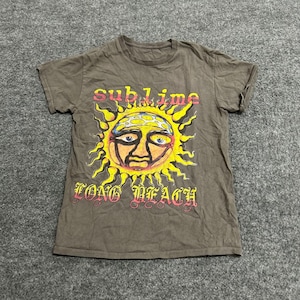 May include: A brown short-sleeve t-shirt featuring a graphic of a yellow sun with a face and blue patterned details. The text "sublime" is printed in red and yellow above the sun, and "LONG BEACH" is printed in red and yellow below the sun.
