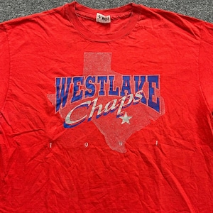 May include: A bright red short-sleeved t-shirt featuring a graphic of the state of Texas in a dotted pattern. The text WESTLAKE is printed in blue block letters, with the word Chaps in a white script font and a small white star below.
