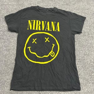 Puede incluir: Una camiseta negra de manga corta con la palabra NIRVANA impresa en letras mayúsculas amarillas en el pecho. Debajo del texto hay un gráfico grande de color amarillo de una cara sonriente con ojos en forma de X y la lengua fuera.