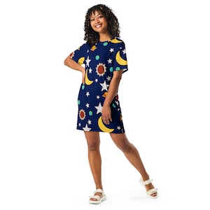 Op de afbeelding: Een marineblauwe t-shirtjurk met een herhalend patroon van gele maansikkels, witte sterren, oranje zonnen en kleine groene en blauwe planeten. Het kledingstuk heeft korte mouwen, een ronde halslijn en een rechte zoom die boven de knieën valt.