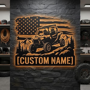 Könnte beinhalten: Ein schwarzes Metall-Wandschild mit einer hinterleuchteten Silhouette eines Geländewagens, der durch eine Berglandschaft mit Tannen fährt, vor einer gealterten amerikanischen Flagge. Der Text [CUSTOM NAME] steht am unteren Rand.