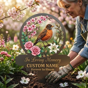 May include: A circular garden memorial stake featuring a brown robin perched on a branch surrounded by pink cherry blossoms, pink roses, and white lilies. The plaque includes the text In Loving Memory, CUSTOM NAME, and Forever In Bloom.