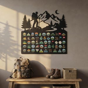 Peut inclure: Un panneau d'affichage mural en métal noir présentant la silhouette d'un randonneur, des montagnes, des pins et un croissant de lune. La base rectangulaire est recouverte de nombreux petits écussons colorés aux formes variées.