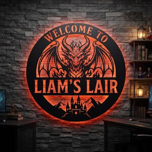 May include: A circular black metal wall sign featuring a backlit orange glow, a central dragon illustration, and the text "WELCOME TO LIAM'S LAIR" above a silhouette of a castle and mountains, mounted on a dark gray stone-textured wall.