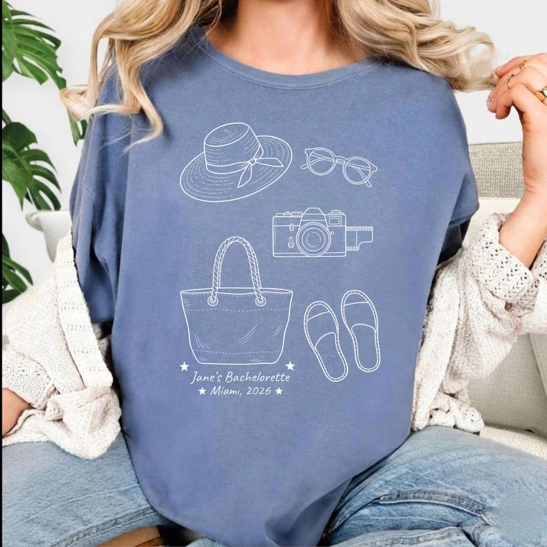 May include: A blue crewneck sweatshirt featuring white line art of a sun hat, sunglasses, a camera, a tote bag, and sandals. Below the illustrations, the text reads: Jane's Bachelorette, Miami, 2026, flanked by two small stars on each side.