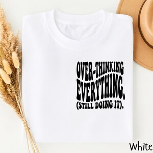 May include: A white t-shirt featuring black text on the chest that reads OVER-THINKING EVERYTHING. (STILL DOING IT). The shirt has a crew neckline and short sleeves, presented in a clean, straightforward layout for clear visibility of the graphic.