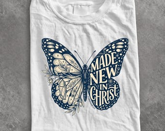 May include: A white t-shirt featuring a large butterfly graphic in dark blue and cream. The left wing displays floral patterns and a chrysalis, while the right wing contains the text "MADE NEW IN CHRIST" and "2 CORINTHIANS 5:17" in a script font.