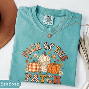 May include: A seafoam green t-shirt with a graphic print featuring three pumpkins and the text "PICK OF THE PATCH". The pumpkins have various patterns including plaid, floral, and solid orange. The text is styled with leopard and checkerboard prints.