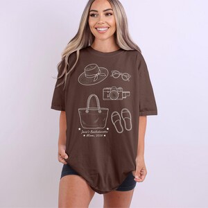 May include: A brown short-sleeve t-shirt featuring white line art illustrations of a sun hat, sunglasses, a camera, a tote bag, and sandals. Below the graphics, the text reads: Jane's Bachelorette Miami, 2026, accented with small stars.