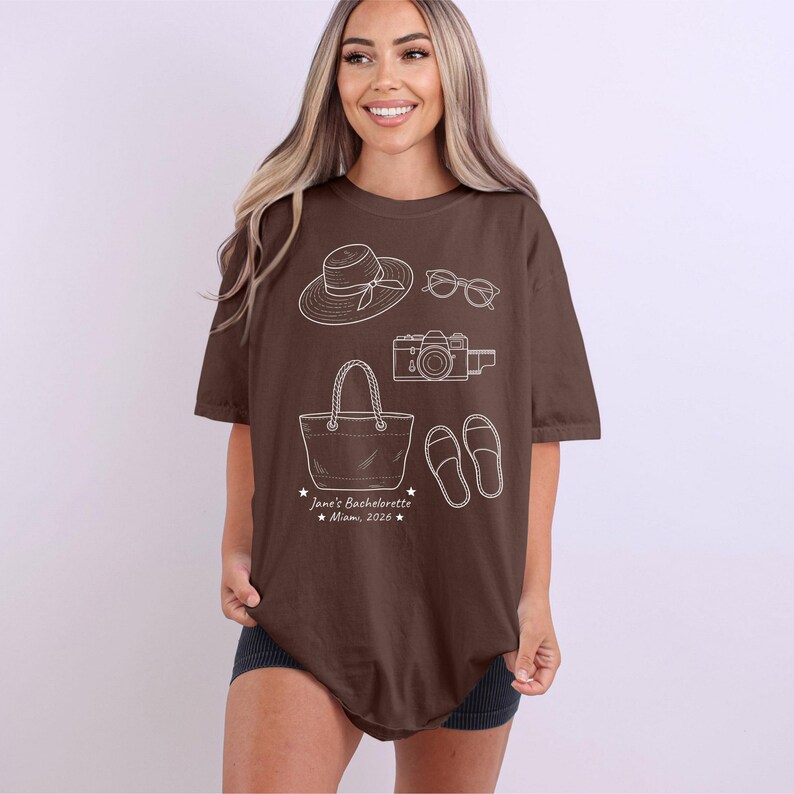 May include: A brown short-sleeve t-shirt featuring white line art illustrations of a sun hat, sunglasses, a camera, a tote bag, and sandals. Below the graphics, the text reads: Jane's Bachelorette Miami, 2026, accented with small stars.